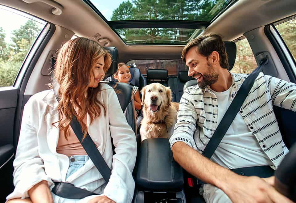 A family of three smiling and looking at their panting dog inside their car