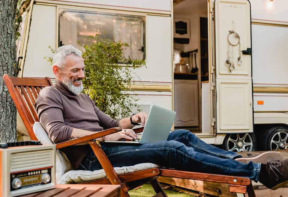 A person sitting on a lounge chair and smiling with their laptop on their lap outside of an RV