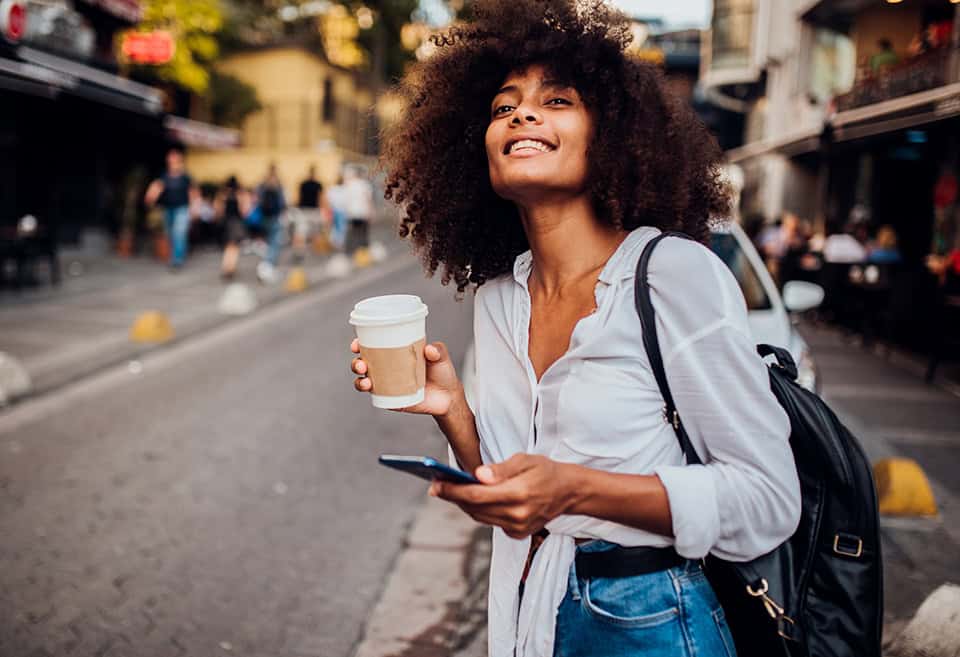 A young person standing on the side of the road holding a cup of coffee and their phone