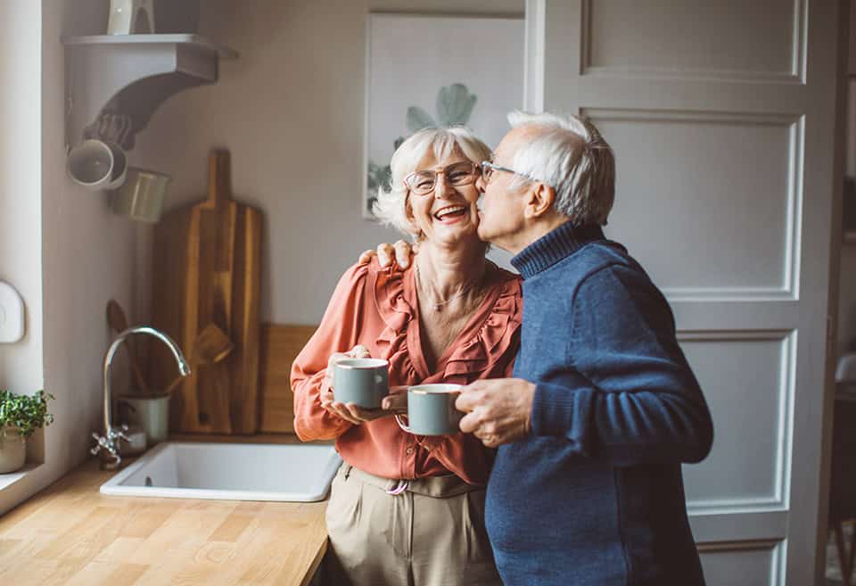 An older couple laughing and hugging while drinking coffee in their kitchen