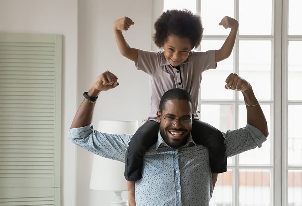 A parent with their child on their back as they both flex their biceps and smile for the camera