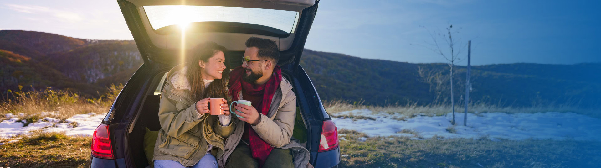 A couple sits in the trunk of their car together outdoors in the snow, clinking their two mugs together filled with hot chocolate.