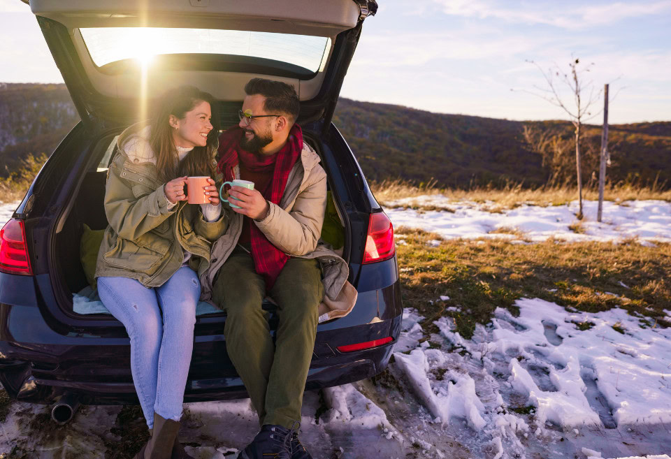A couple sits in the trunk of their car together outdoors in the snow, clinking their two mugs together filled with hot chocolate.