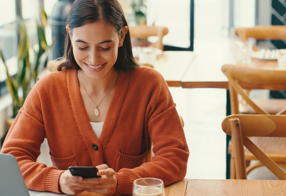 Woman in cafe looking down at her phone, smiling. She's browsing BluPeak's new digital banking features.