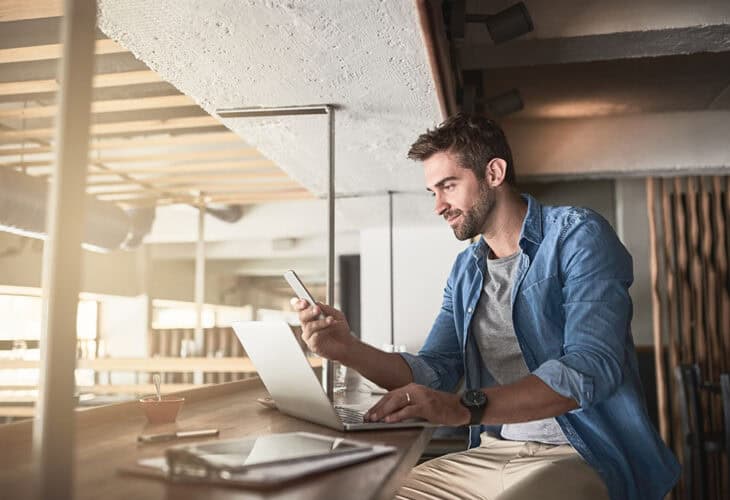 Man sitting at table with laptop open while scrolling through phone