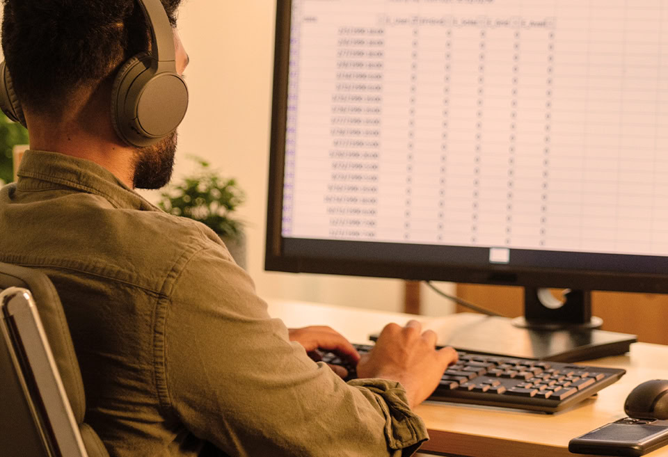Man wearing headphones working at a desktop computer screen with spreadsheet on the display