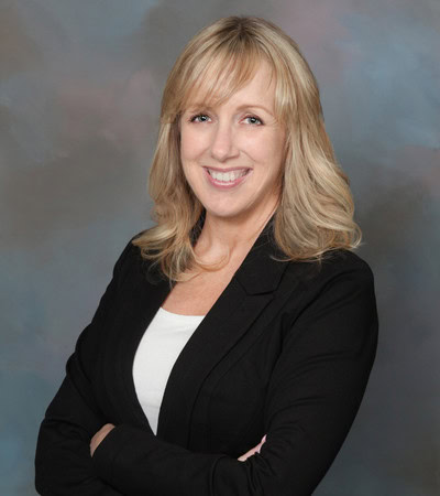 Shelley Duckart, Mortgage Specialist. In the photo, she has blonde, shoulder-length hair and is smiling confidently. She is wearing a black blazer over a white top and has her arms crossed. The background is a soft blend of muted blue, gray, and pink tones, giving the image a formal, studio portrait feel.
